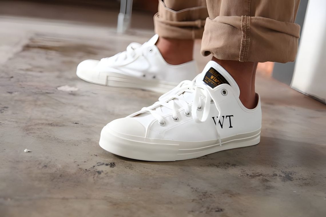 WTAPS white canvas low-top sneakers with 'WT' monogram on toe, worn with cuffed tan trousers.