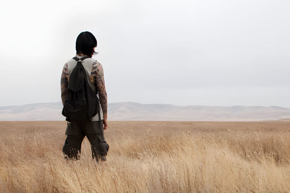 Tattooed male model seen from behind in dry grass field, dark cargo pants and canvas rucksack, Odyn Vovk lookbook.
