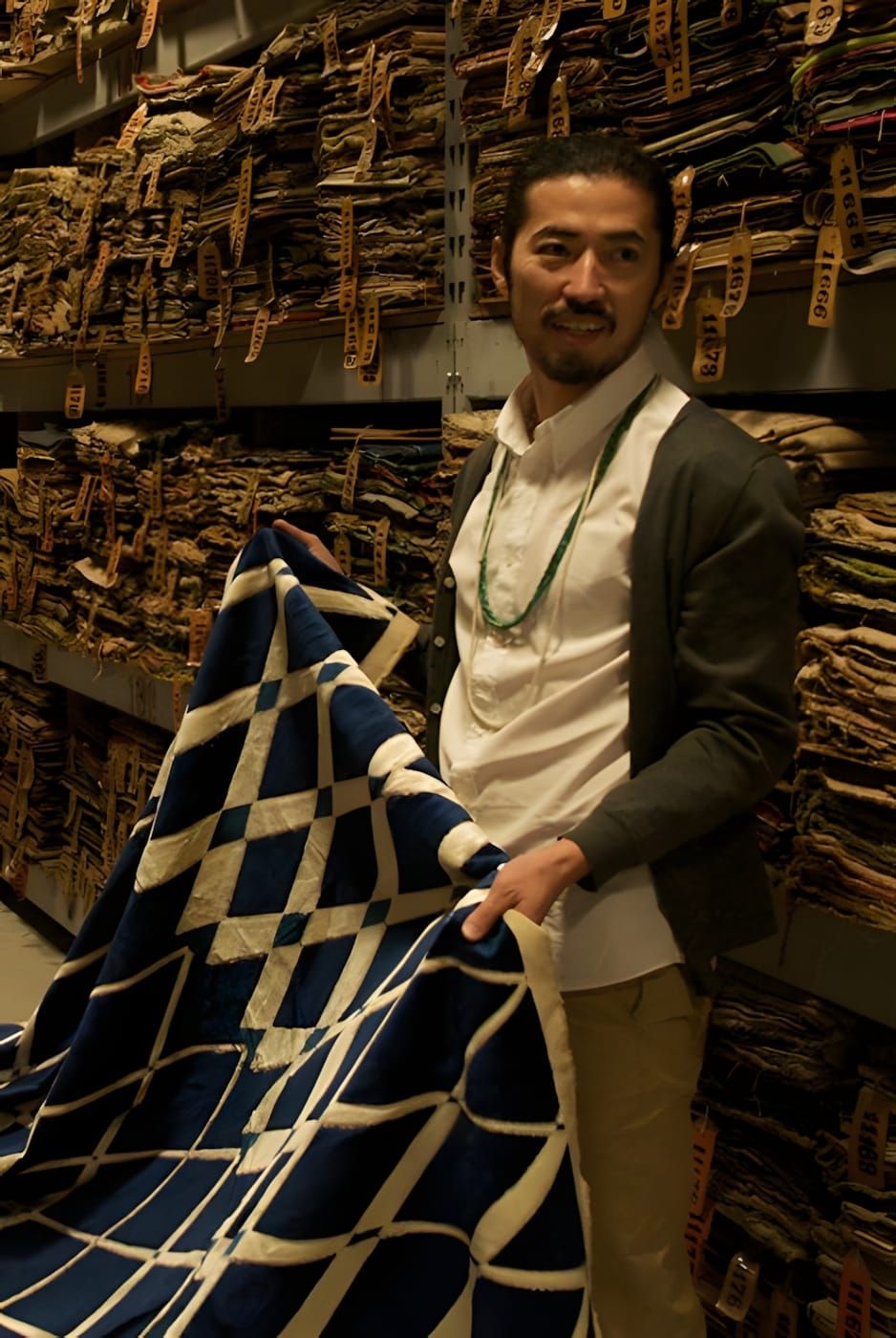 Hiroki Nakamura holds an indigo and cream diamond-pattern patchwork textile in a warehouse lined with tagged vintage fabric b