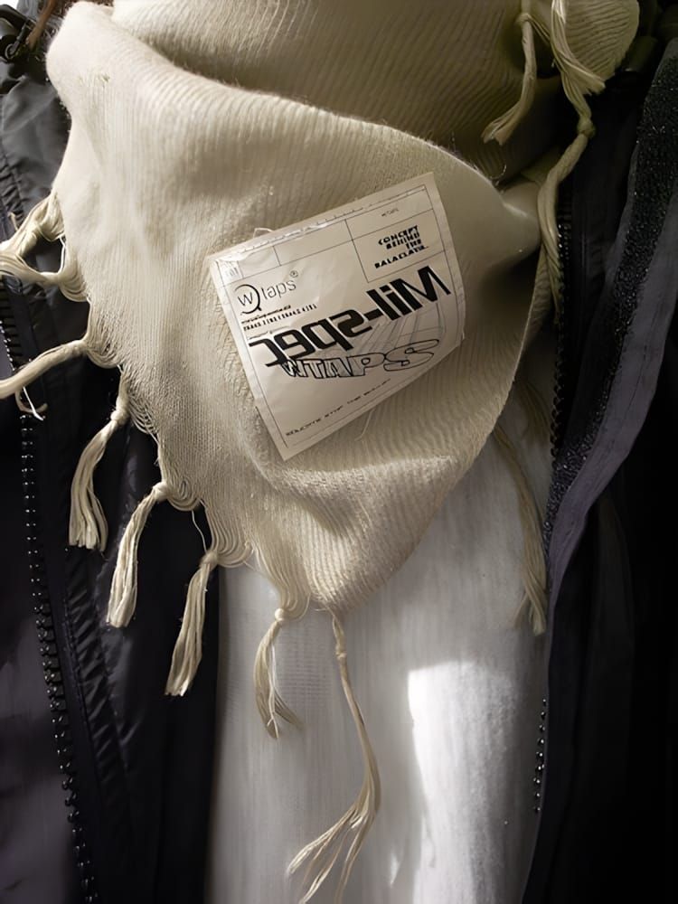 Cream WTAPS Mil-spec shemagh with fringe draped over a dark jacket, WTAPS woven label visible.