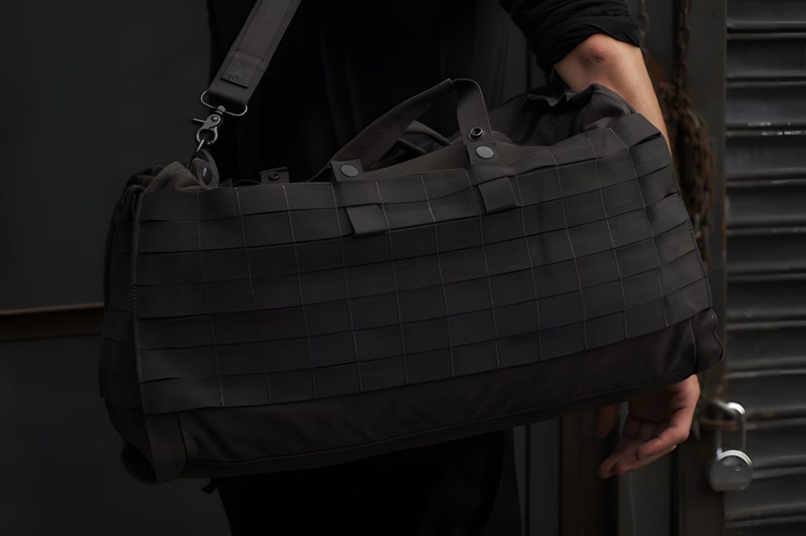 Black barrel duffel bag with MOLLE webbing panel, top handles, and shoulder strap, held by a person outdoors.