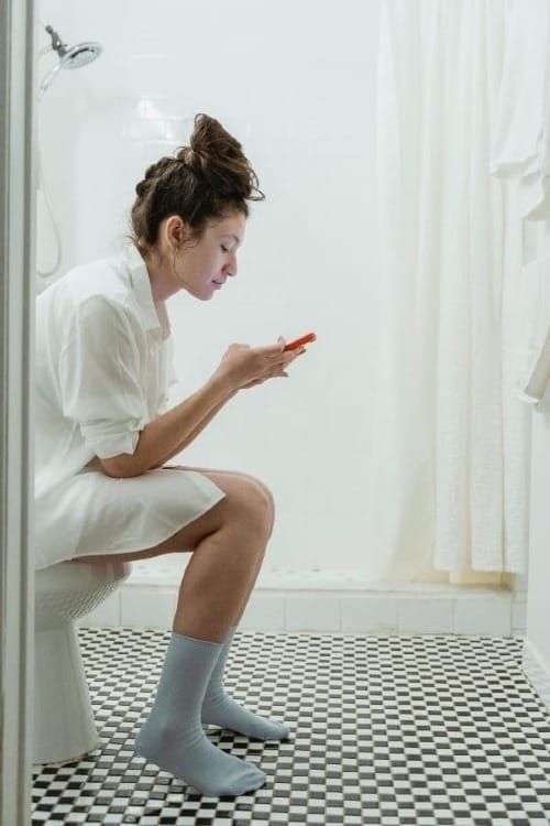 Junge FRau sitzt auf Toilette und guckt in ihr Handy