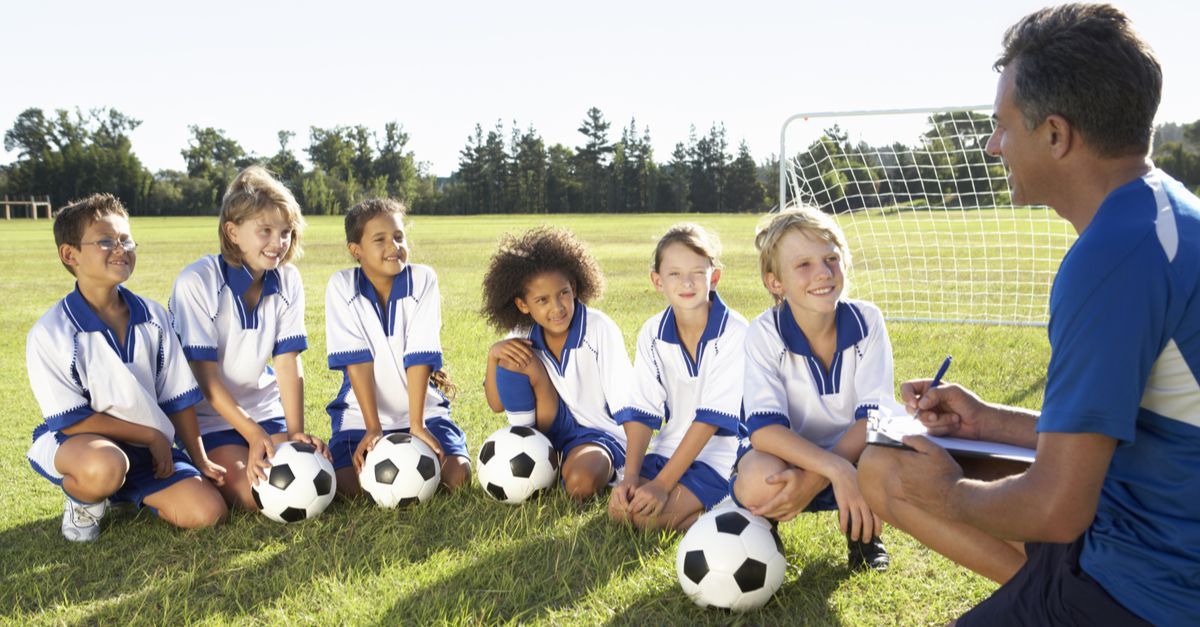 A football coach kneeling on a football pitch talking to a group of six children