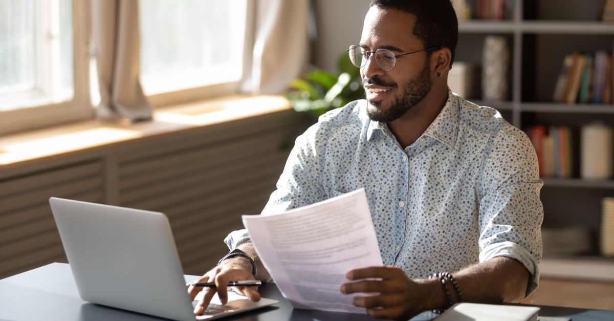A man, half-smiling, typing on a laptop computer and holding some printed sheets of paper