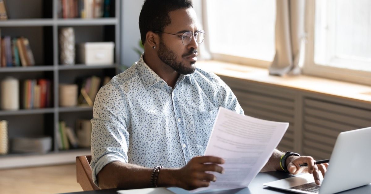 A man sat at a laptop, typing with his left hand and holding some paperwork in his right hand