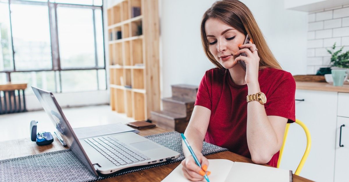 A woman sat at a laptop in a home kitchen, on the phone and making notes in a notebook