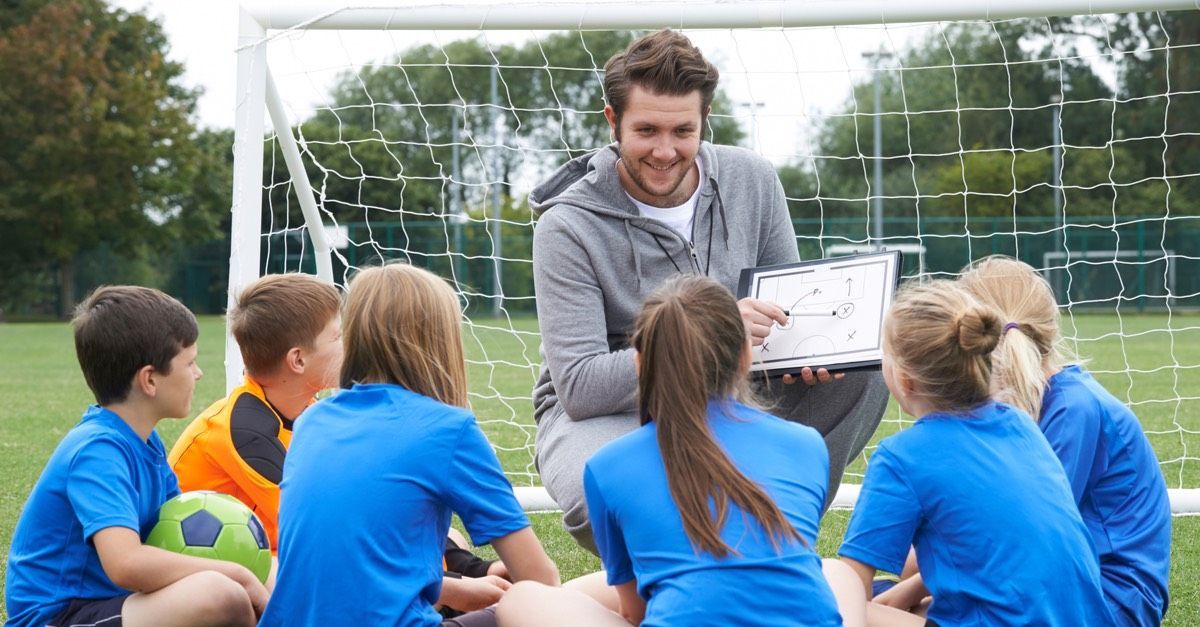 A football coach kneels down to talk to a group of mixed sex elementary school players
