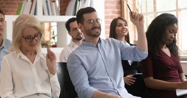 A small group of volunteers sat down in a room. Two of them have their hands raised.