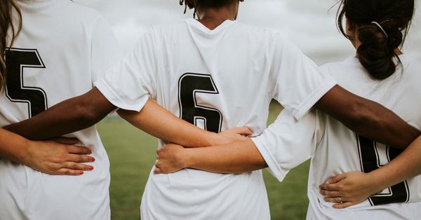 Three female footballers of different races face away from the camera, with their arms around each other's waists