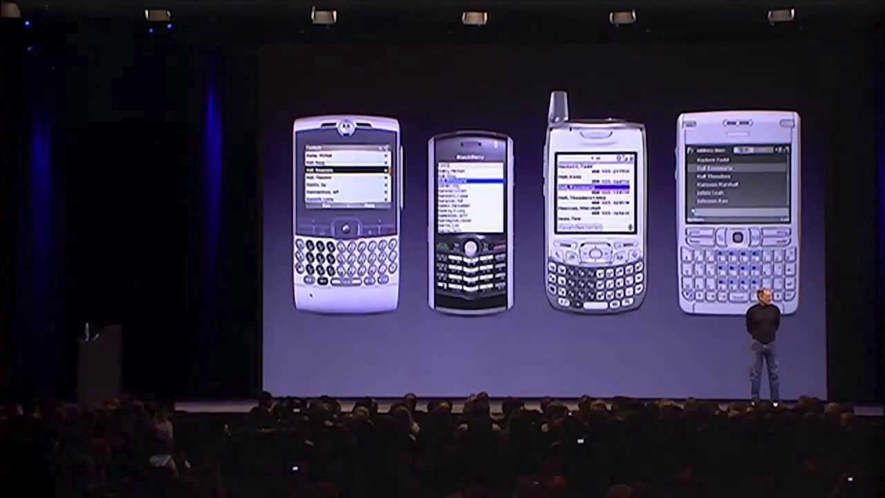Steve Jobs on stage at the 2007 iPhone launch in front of a slide showing competing smartphones including the Palm Treo and BlackBerry