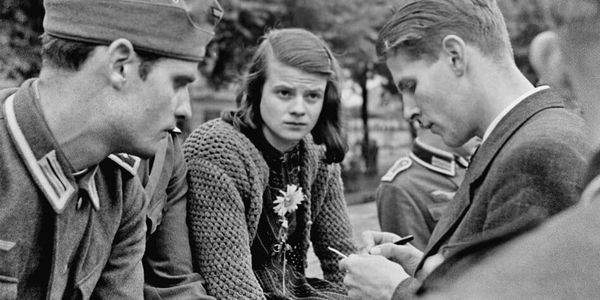 A photo of Sophie Scholl, her brother Hans in a Wehrmacht uniform and Christoph Probst who is writing on a notepad. She is looking at Christoph and has a flower pinned to her clothing.