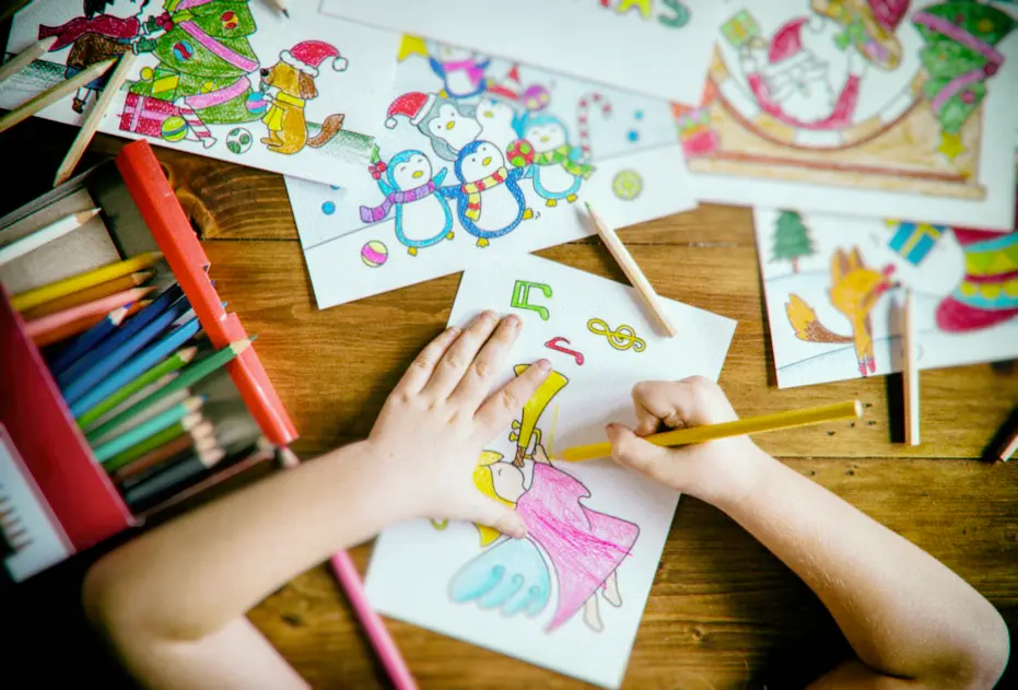 un enfant dessinant aux crayons de couleurs sur des feuilles de papier posées sur une  table en bois