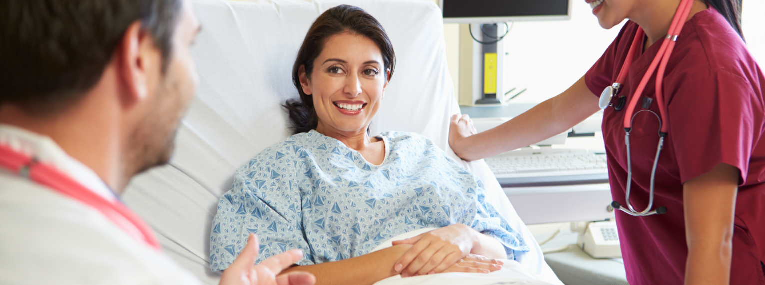 Eine Patientien liegt in einem Krankenhausbett und spricht mit einem Arzt und einer Krankenschwester.