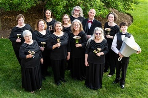 Group of 11 women and 1 man dressed in black, standing together on a grassy surface holding individual handbells and looking up toward the camera.