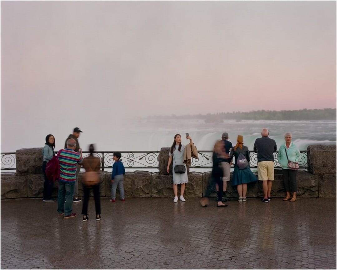 The image shows a group of people standing on a paved surface with a stone and wrought iron railing nearby. In the background, there is a misty waterfall merging into a pink sky, with green trees in the distance. Some individuals appear blurry due to motion. The color scheme includes earthy tones, pastel shades in the sky, and vibrant greens and whites from the natural elements.