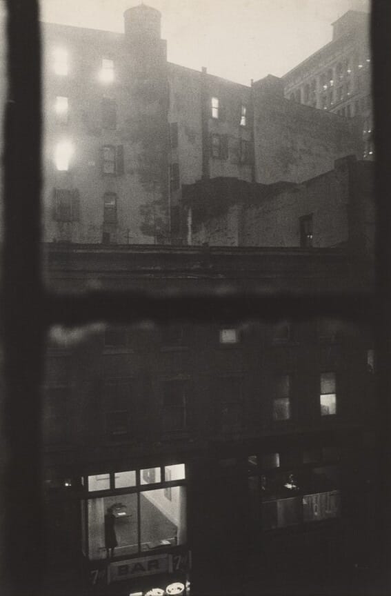 The image shows an urban scene from an elevated viewpoint, looking out from a window across a row of buildings. The skyline consists of building tops, with a larger distant building visible in the background. In the middle section, more buildings with dimly lit windows are visible. Near the foreground, a bar with a figure inside is seen through large windows. The image is monochromatic with a grayscale palette, conveying a somber mood, heightened by the contrast between dark buildings and illuminated windows. The image appears to use a soft focus or blurred technique.