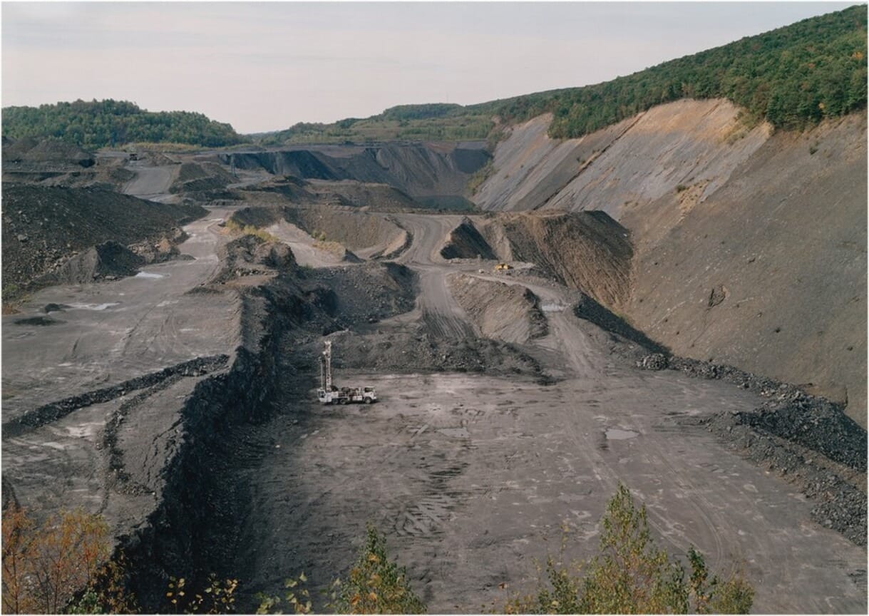 The image depicts a mining site with a drilling machine next to a large open pit. The surrounding landscape is mostly gray and brown with patches of green vegetation. In the background, there are hills covered with greenery, separating the land from the sky. The terrain shows tire tracks and piles of excavated material on a dirt track. The colors are muted and earthy, with hints of green contrasting against the primarily gray and brown landscape.