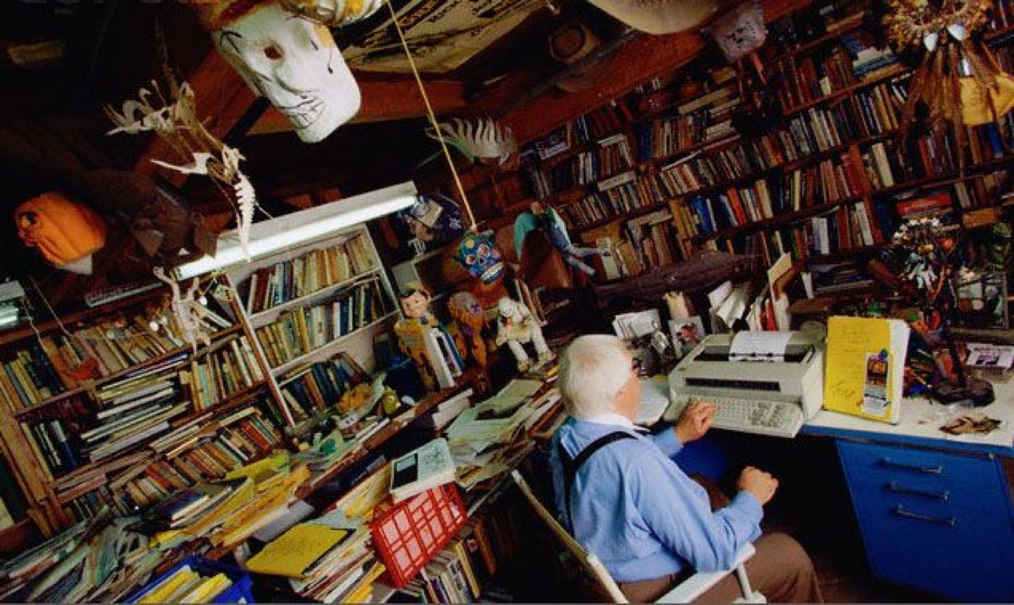 Ray Bradbury sitting at his desk writing, surrounded by books and other objects