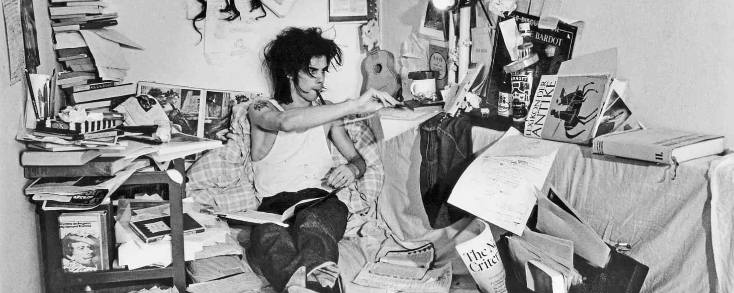 Nick Cave writing in a cluttered area full of books, papers and cushions