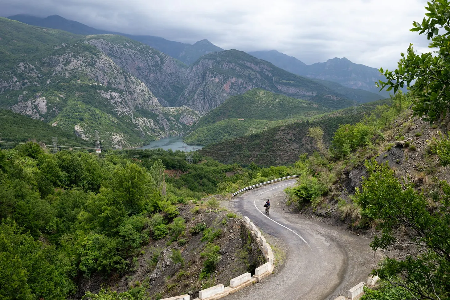 Foreground, a cyclist makes their way through a winding mountain pass, while in the distance a mountainous landscape lies ahead