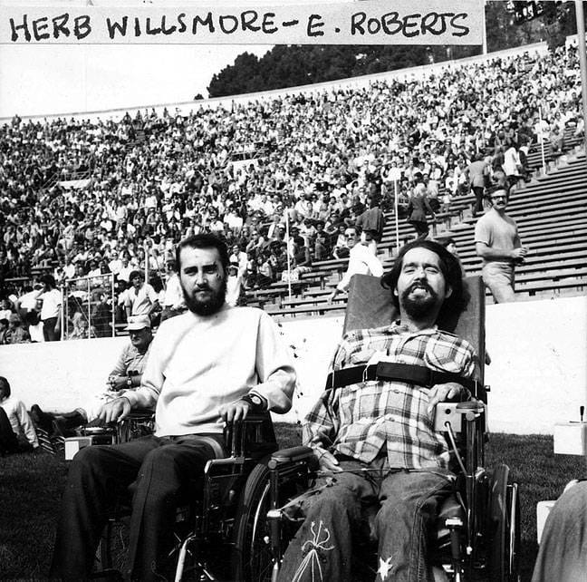 black and white photo, two young men sitting in wheelchairs, large crowd in the bleachers of a college football stadium. text up top that says "Herb Willsmore-E. Roberts"