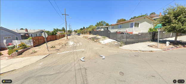 Image of a sloped intersection, looking down the street with no proper sidewalks. Single-story single-family homes and both metal and wooden fences.