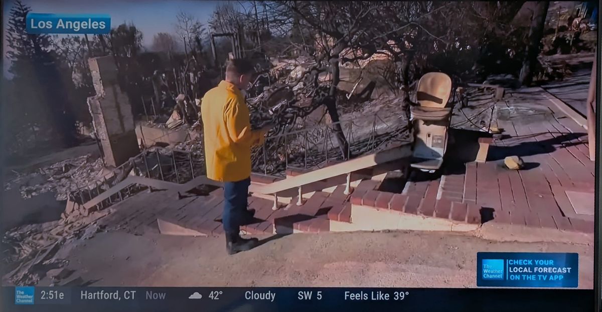 A reporter from the Weather Channel looks at a charred adaptive stair lift going up a brick staircase to a burned-out home
