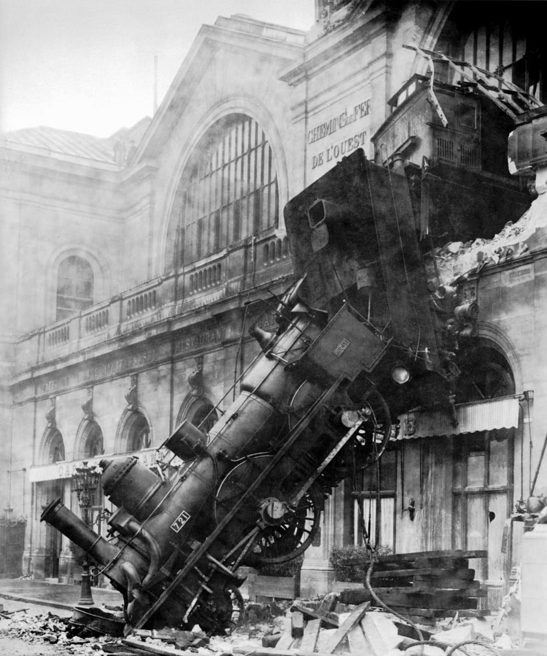 Old black-and-white photo. Steam engine train crashed through the second floor of a two-story building in a city, and is hanging off the edge of the building on to the street.