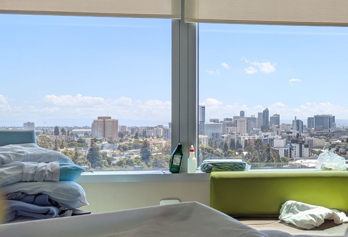 Inside of a hospital room looking out a window with an aerial view of Oakland's downtown. Tree-filled neighborhood nearby, office and residential towers farther away.
