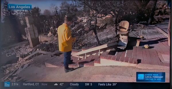 A reporter from the Weather Channel looks at a charred adaptive stair lift going up a brick staircase to a burned-out home