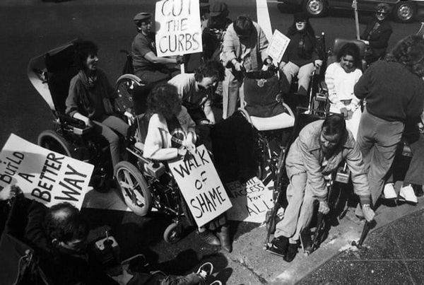 Black-and-white photo. Around a dozen people in old school wheelchairs hold signs. One man with a disability smashes the curb with a hammer. 