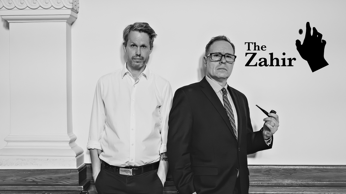 Black-and-white promotional photograph of two men standing side by side. One wears a light button-down shirt; the other wears a dark suit and holds a pipe.