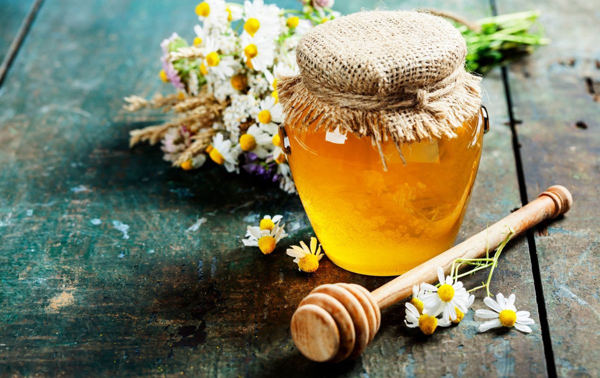 A jar of honey, a honey dipper and chamomile flowers
