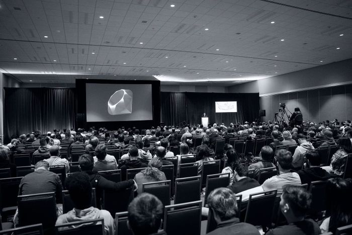A large audience watching a conference talk, with the Ruby gem logo displayed on screen