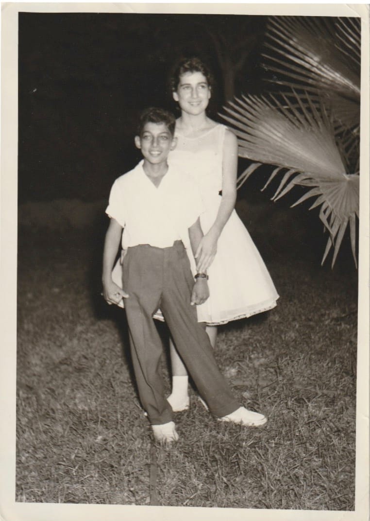 A young girl and her younger brother, posing on grass with a palm behind them