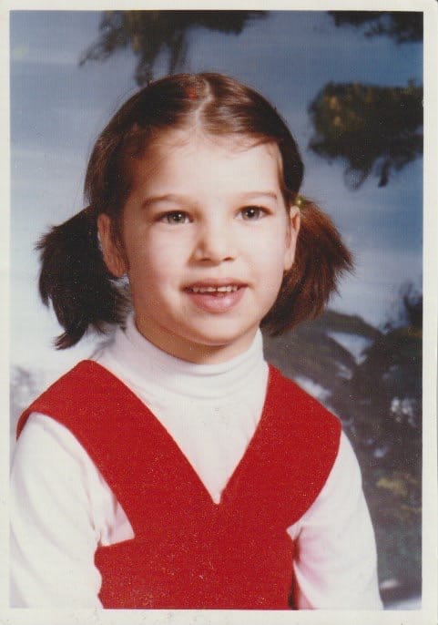 A school photo of young girl with brown hair in pigtails, wearing a white turtleneck under a red corduroy jumper.