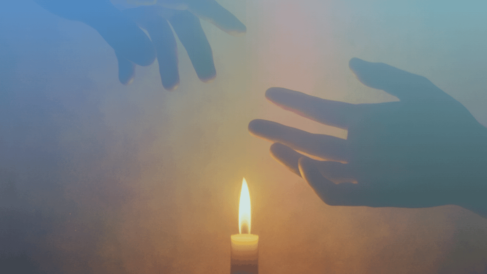 Two hands stretching toward each other but not touching in a room lit by a candle