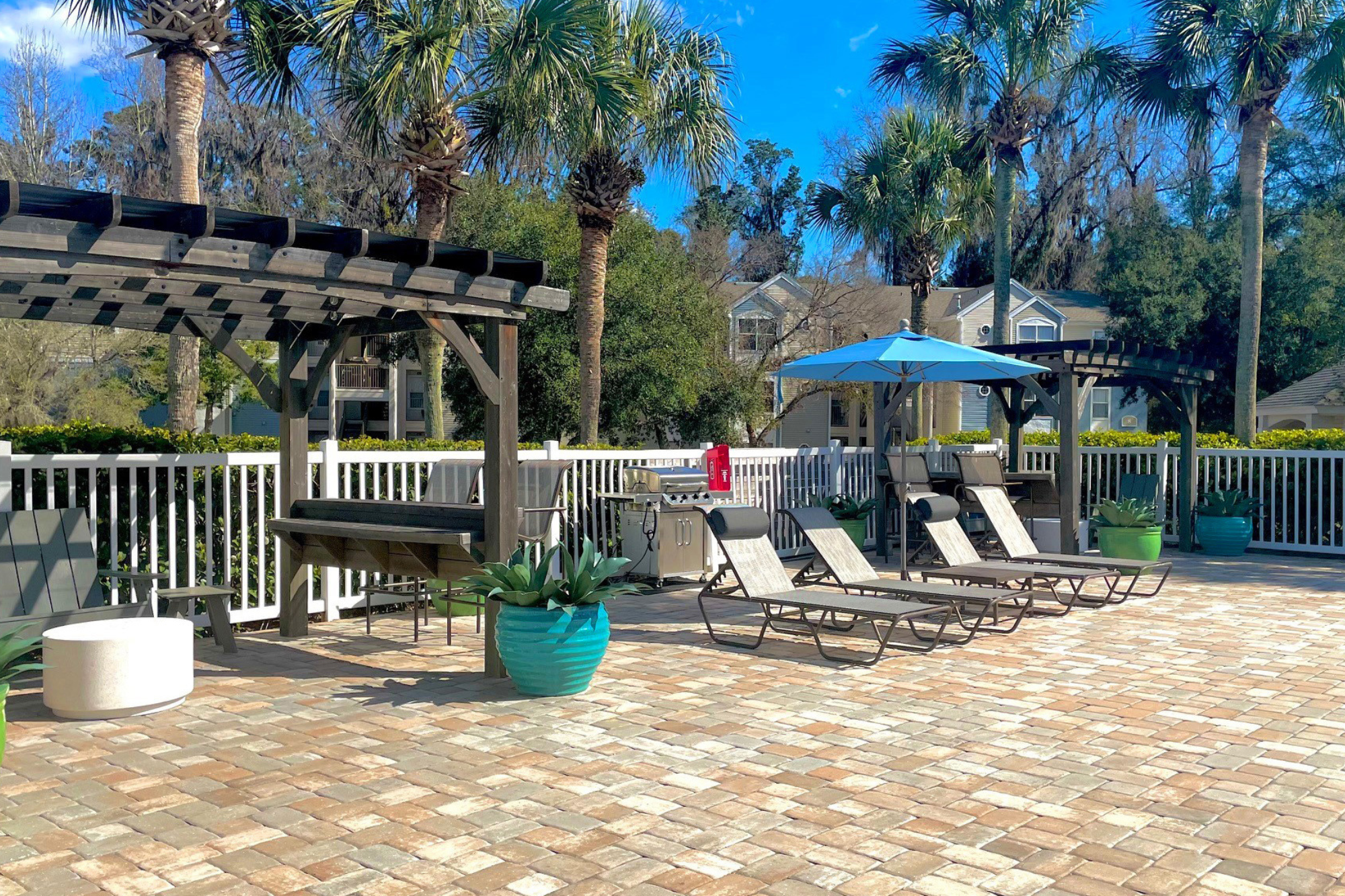 Lounge chairs on a patio receiving sun light, with a gas grill in the background and trees surrounding for a calm feeling