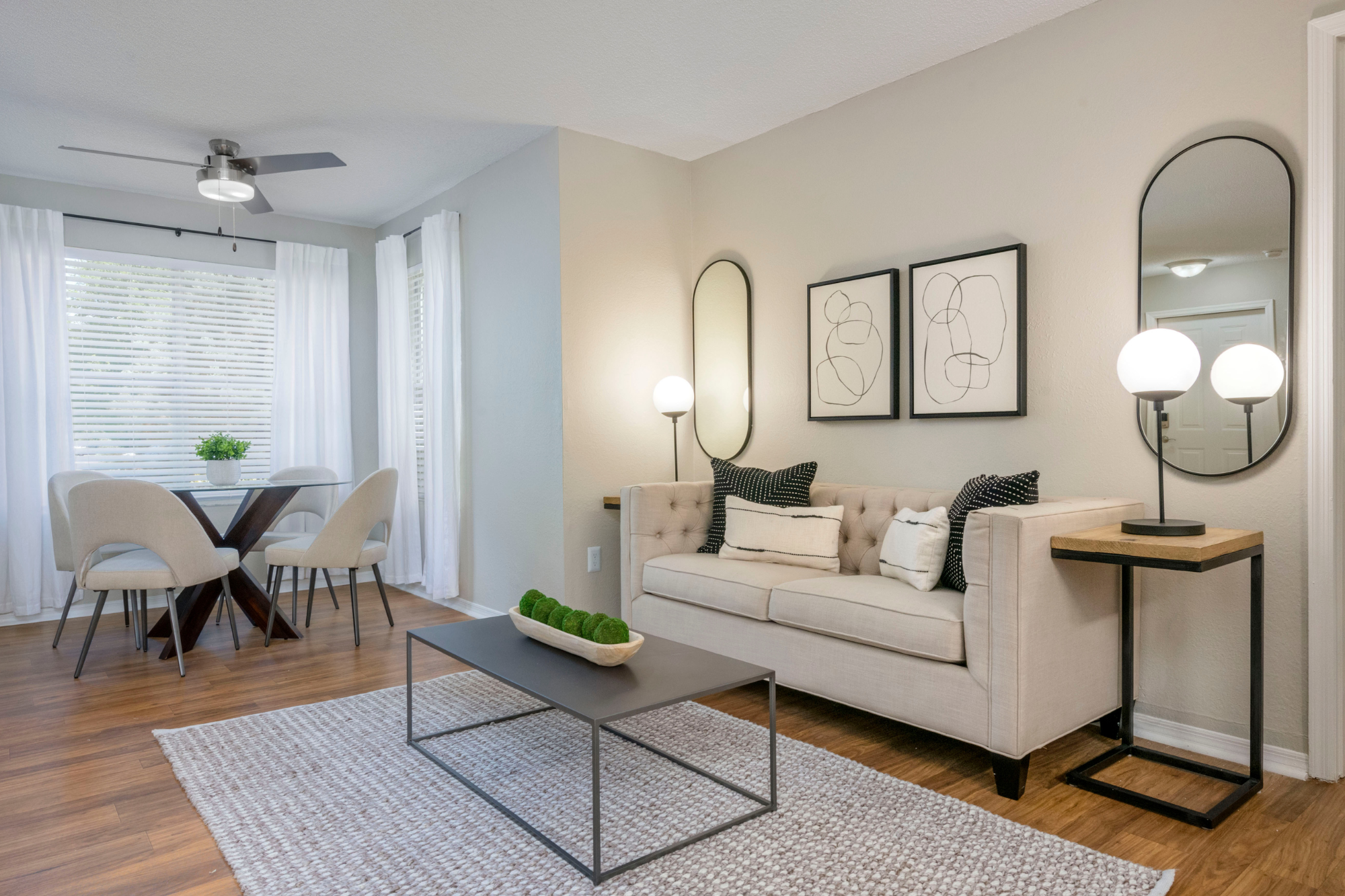 A bright and welcoming living room complete with a cream colored couch and decorative pillows, with mirrors and floor lamps on either side. In the open space is also a nook for a dining room table with a ceiling fan above it.