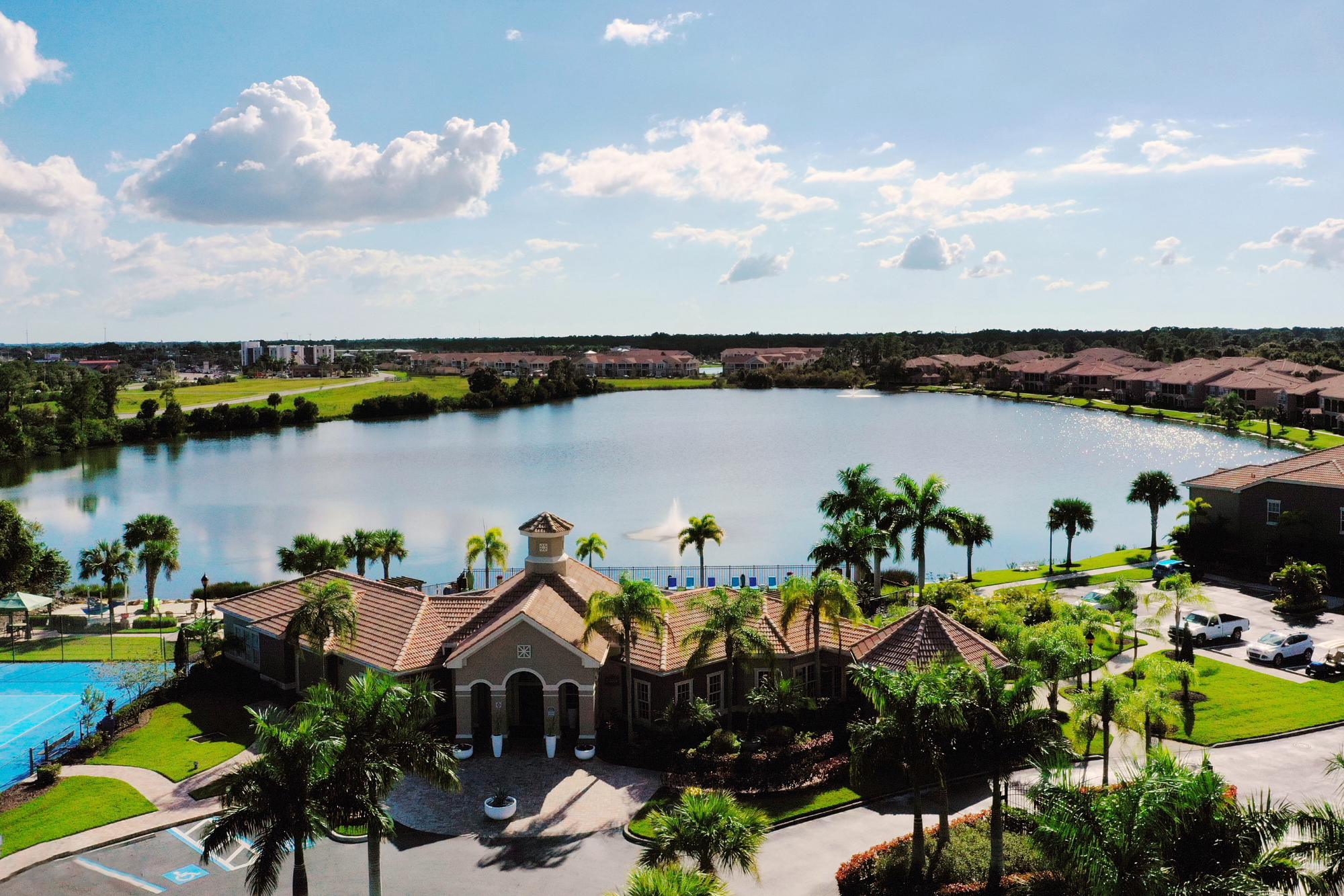 Aerial view of Lakes of Tuscana community, showing numerous apartment buildings circling the lake with a waterfall in the center and beautiful palm trees surrounding.