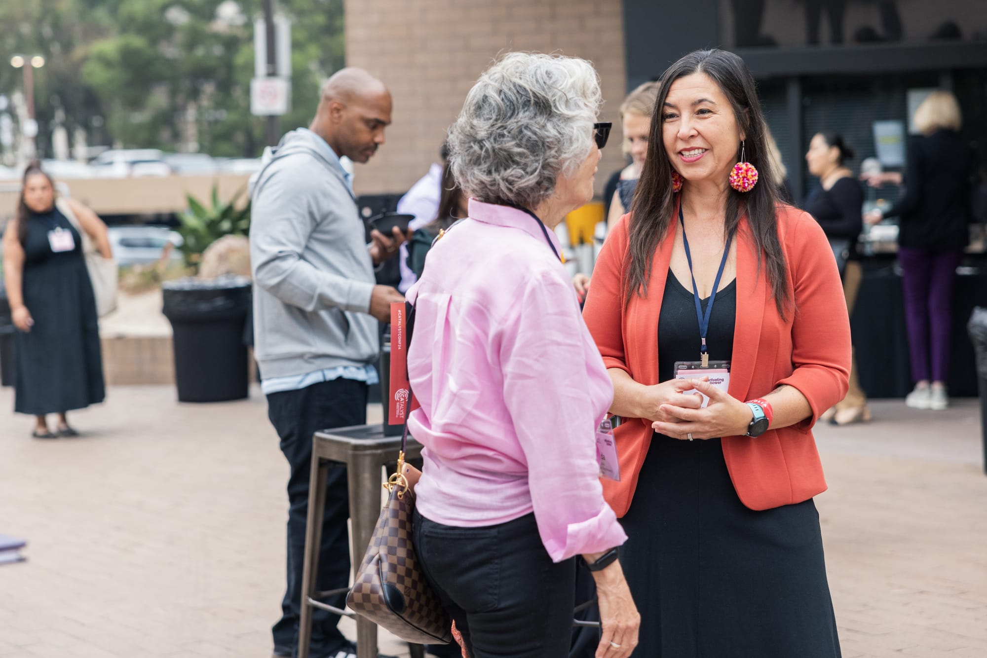 Two women stand together talking outside with other people visible in the background of the networking event