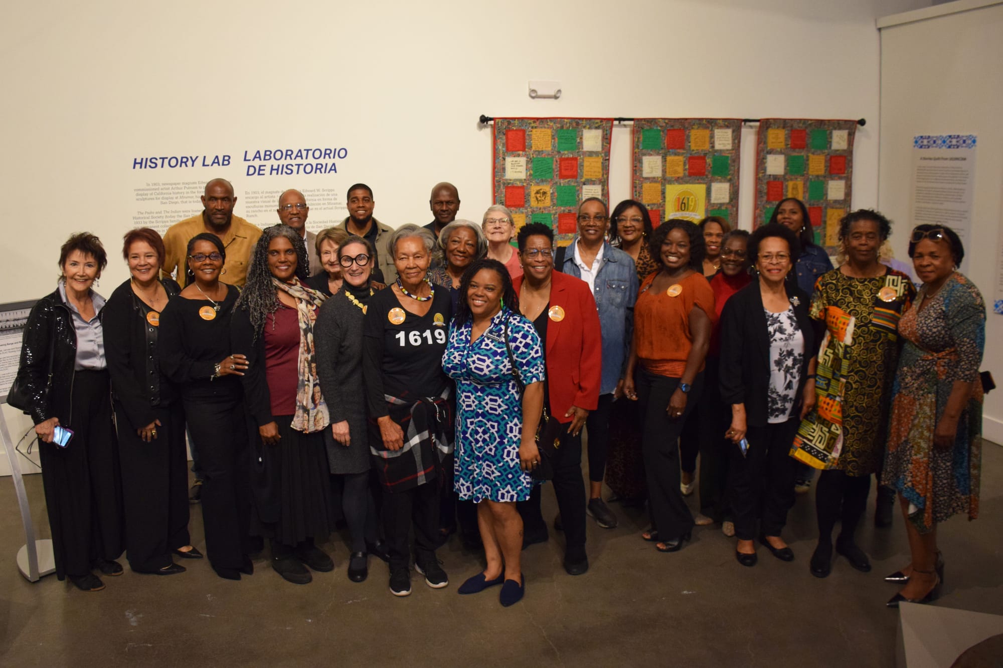 A group of people stand in front of a panel of quilts hung on the wall
