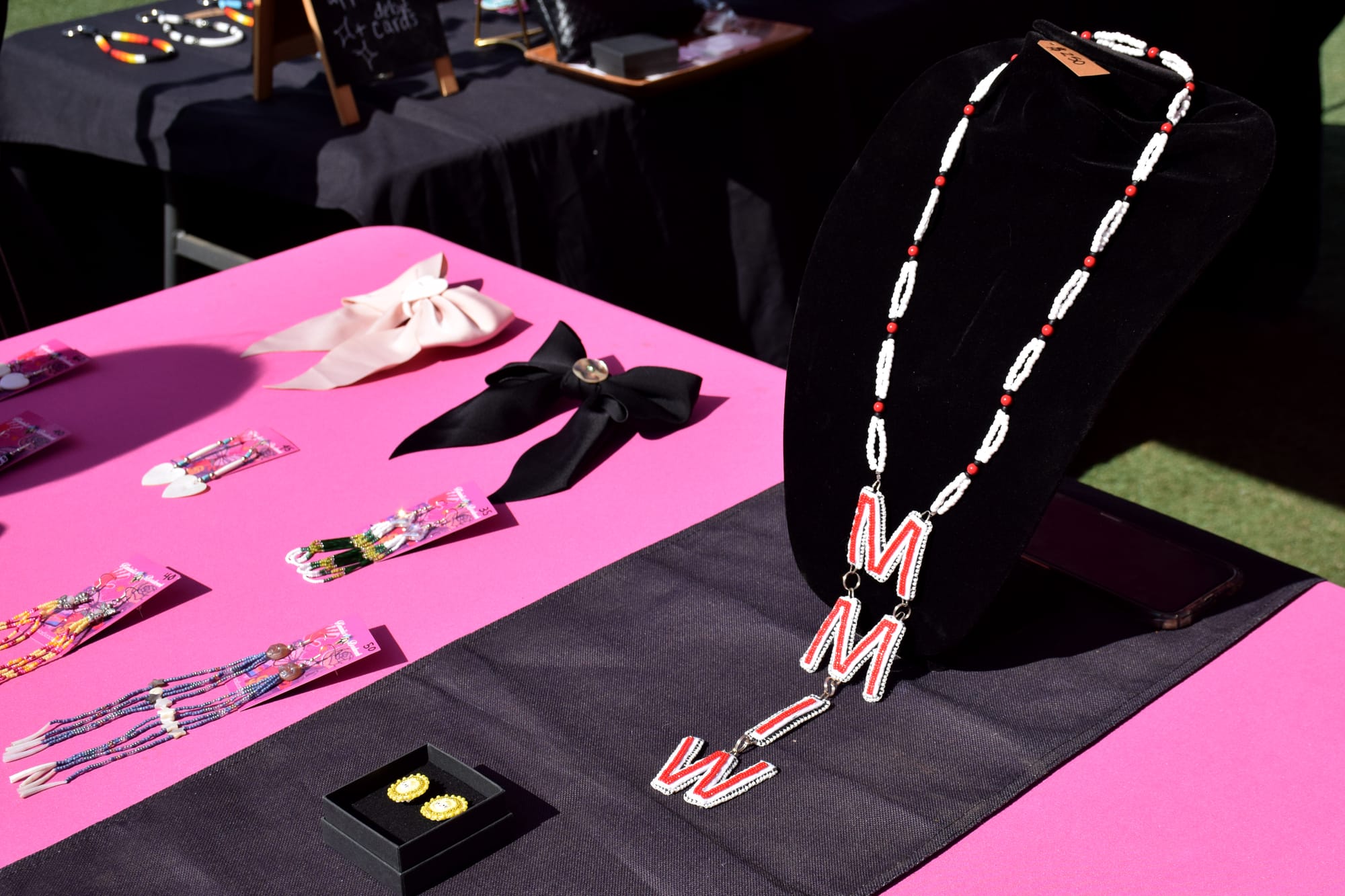 A pink and black table display with earrings, bows and a beaded necklace with the acronym MMIW in red and white beads.