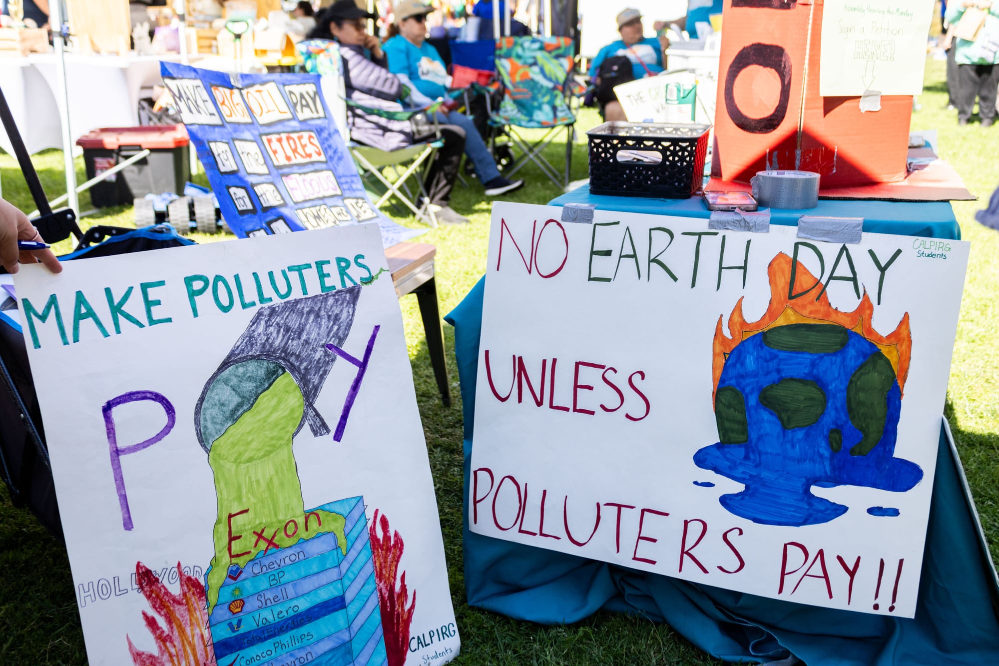 Handmade signs at an outdoor event read "Make Polluters Pay" and "No Earth Day unless polluters pay"