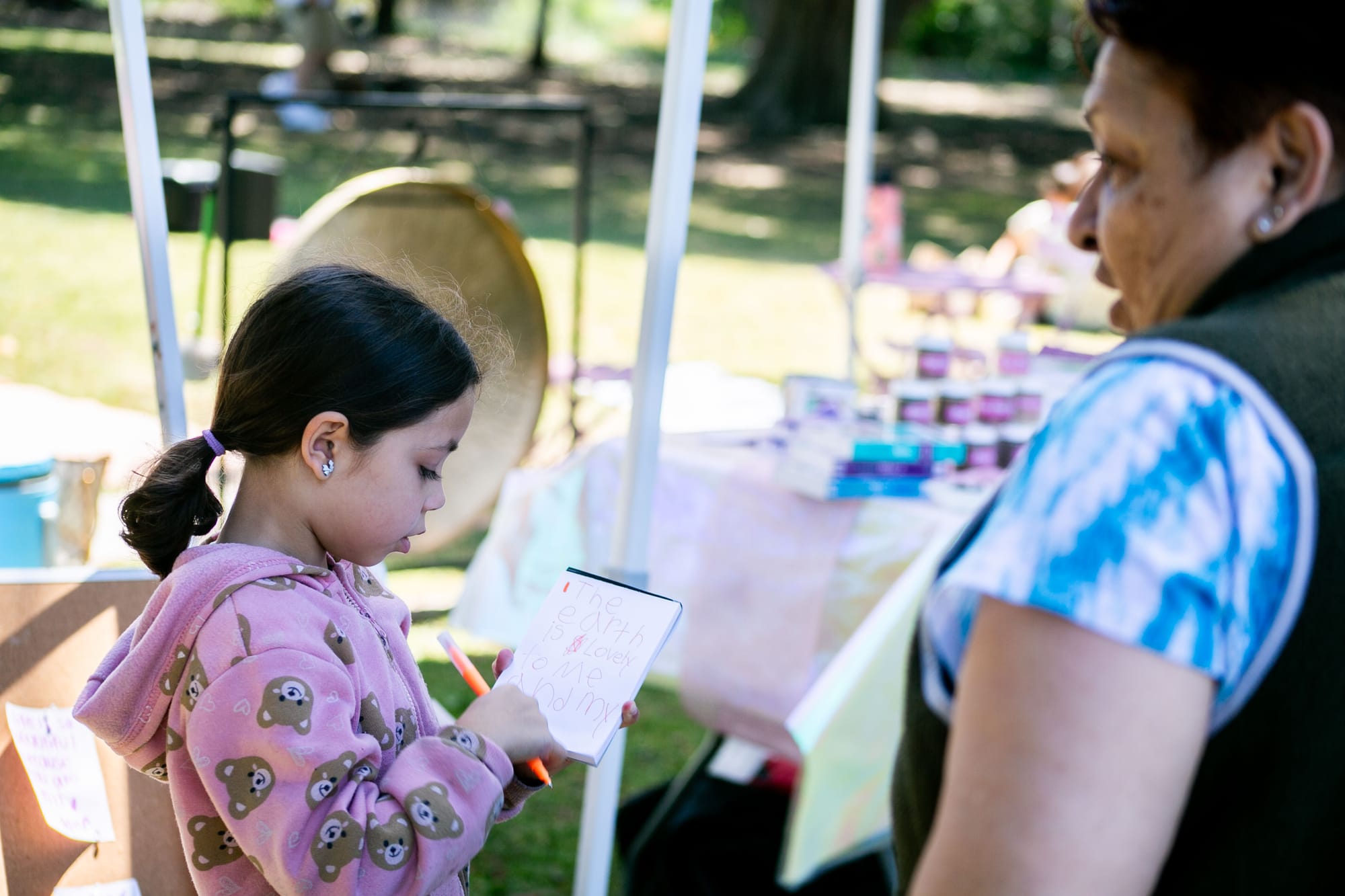A girl in a pink hoodie with brown bears on it writes a note next to her grandmother at an outdoor event