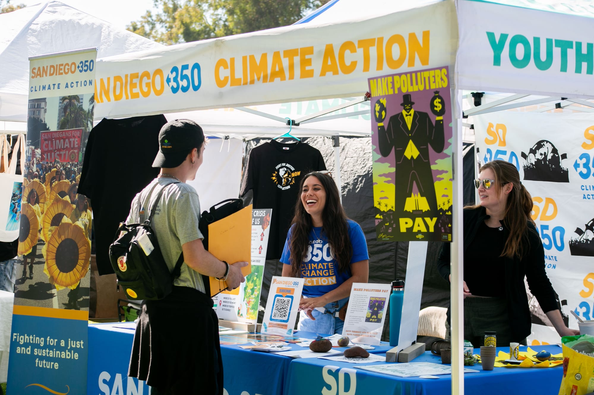 Two women speak to a man at a booth with the words "San Diego 350 Climate Action Youth" and "make Polluters Pay"