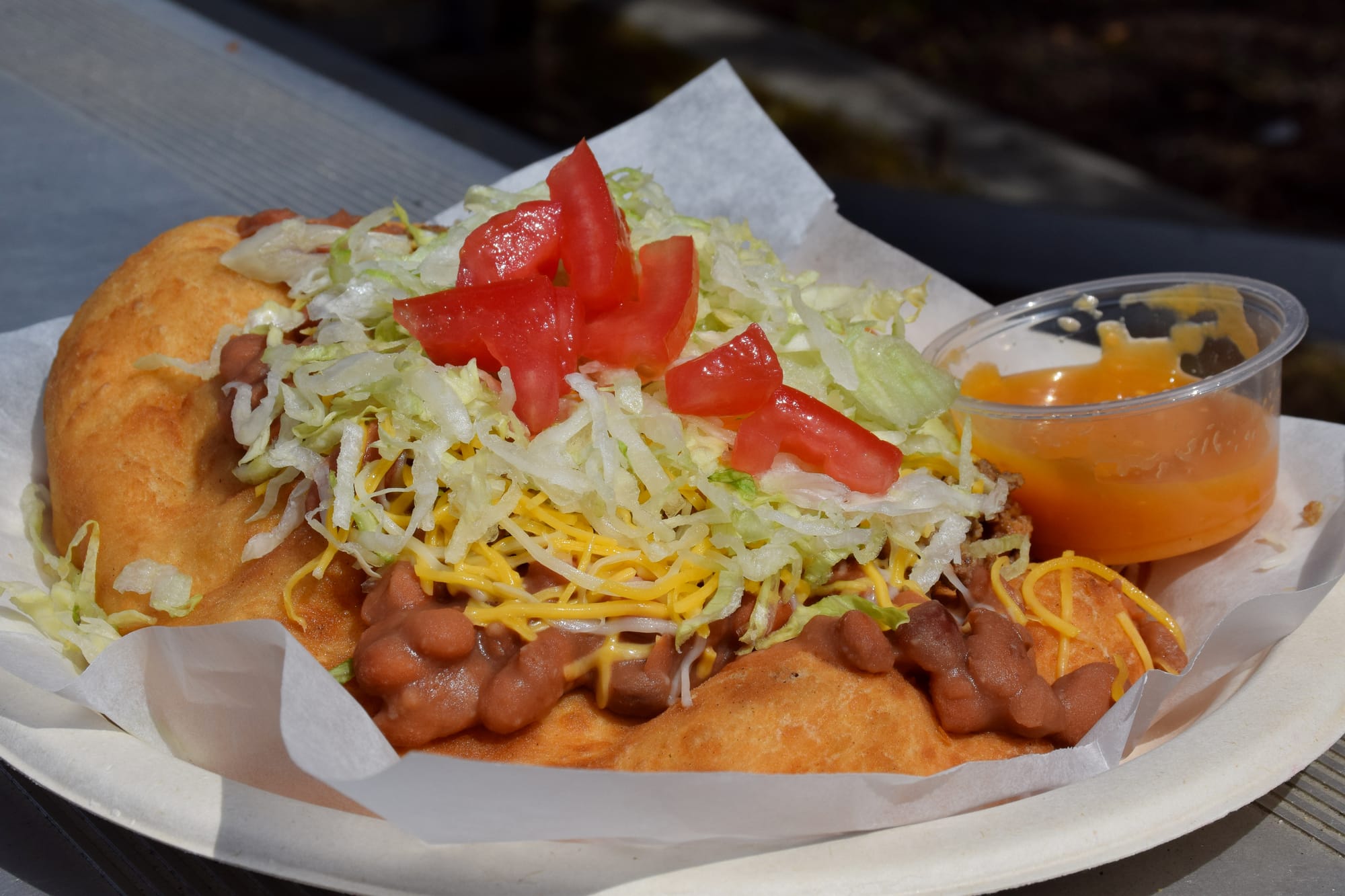 A piece of frybread on a paper plate, topped with beans, cheese, lettuce and tomatoes with a cup of salsa on the side.