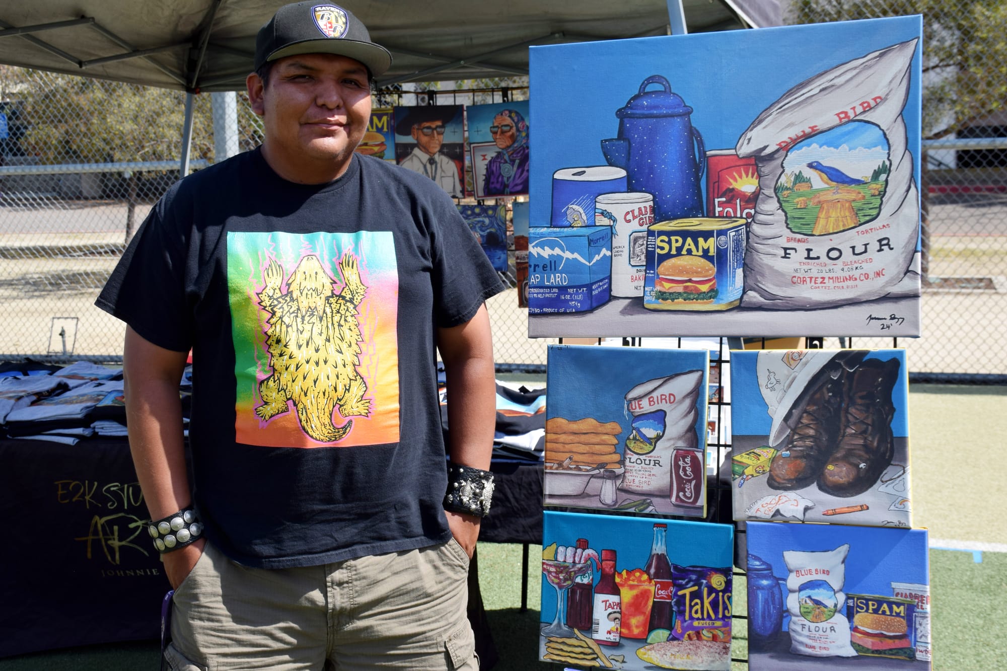 A man poses for a photo next to a display of still art paintings featuring items like Bklue. Bird Flour, Spam and Takis.