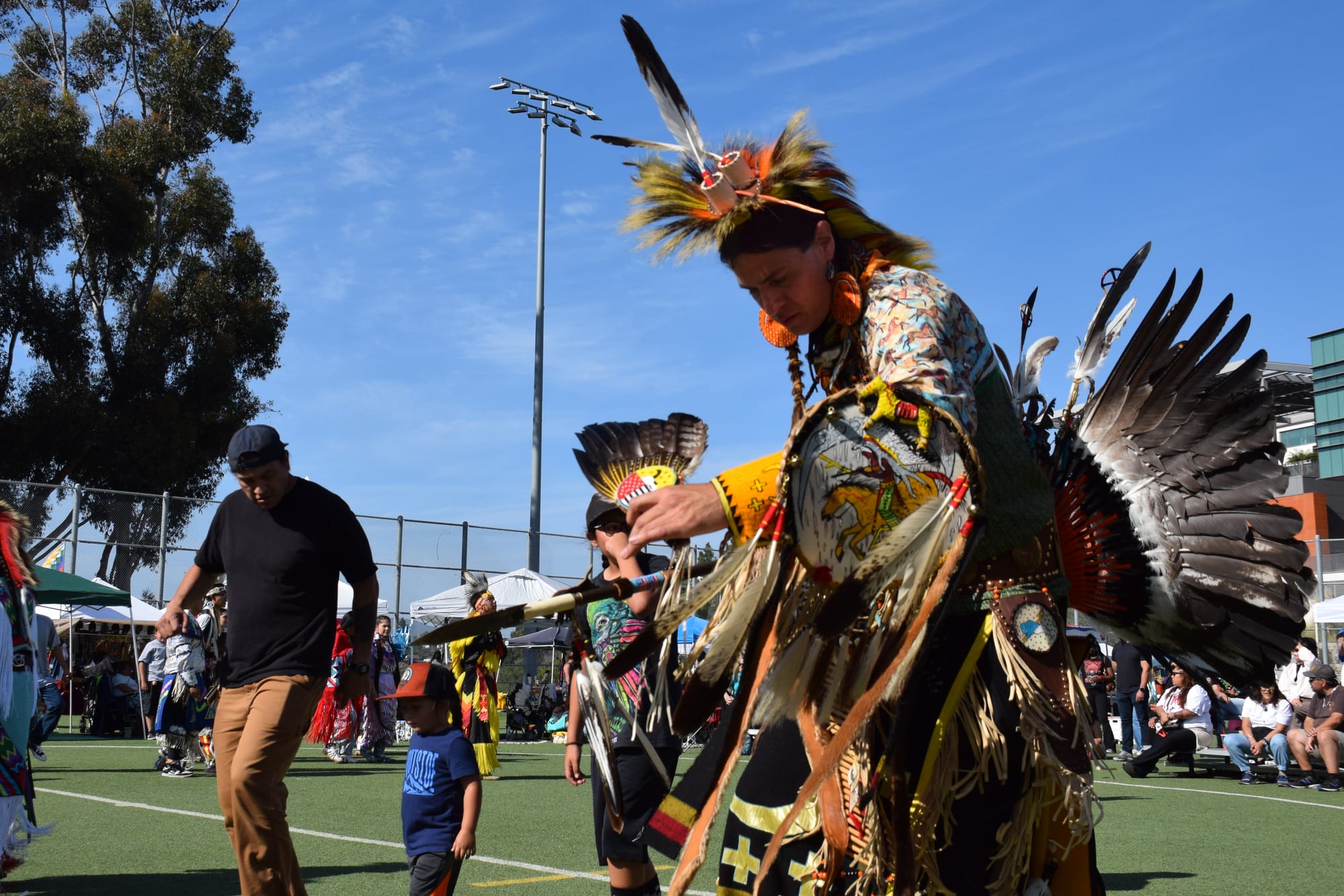 A man wearing a traditional regalia and a bustle made up of feathers dances outside at a powwow.