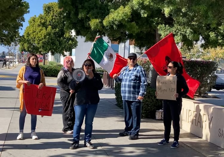 Five people holding flags, signs and a megaphone stand outside a library on a sidewalk.
